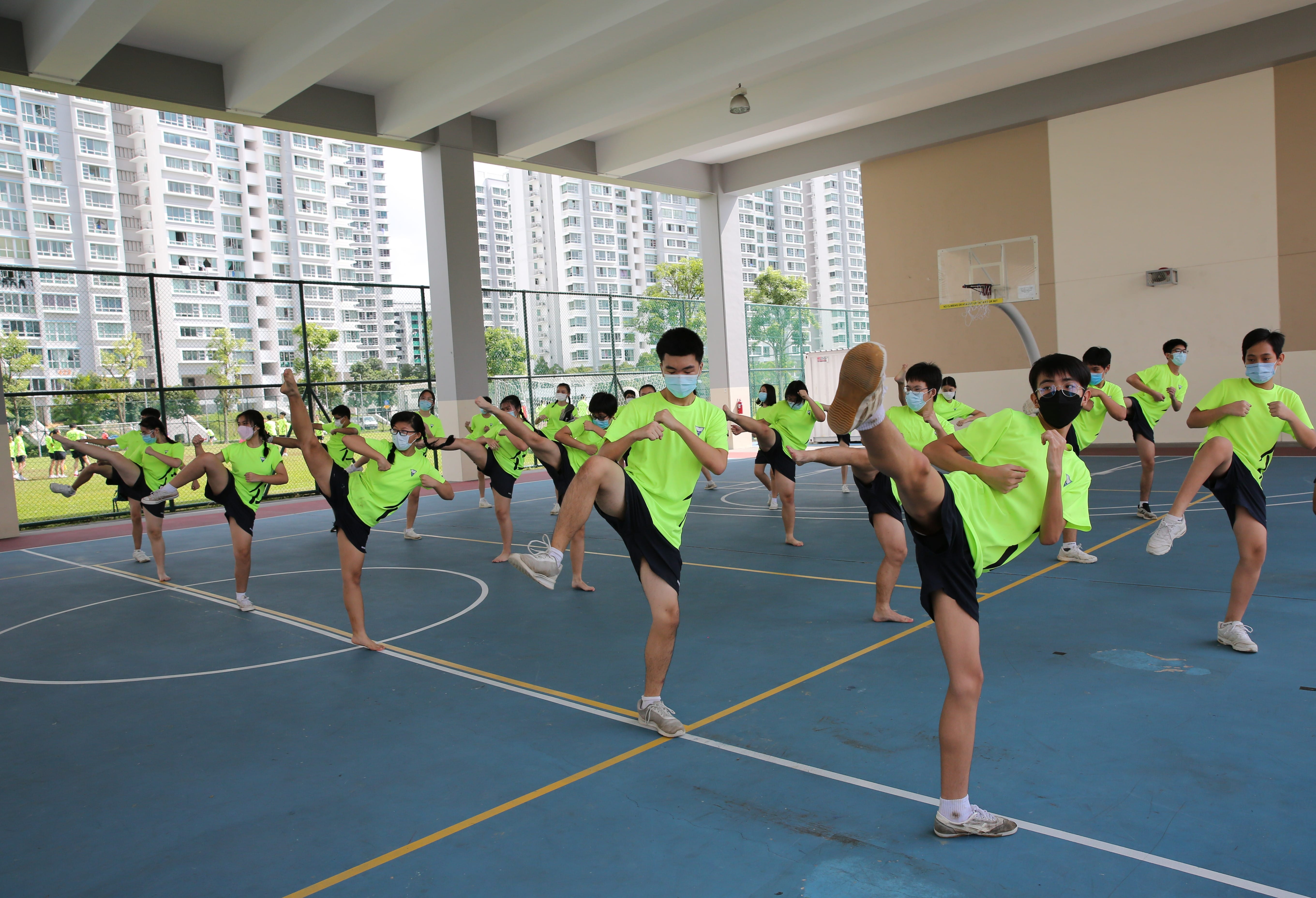 Students in green shirts and masks perform synchronized high kicks on an outdoor covered basketball court.